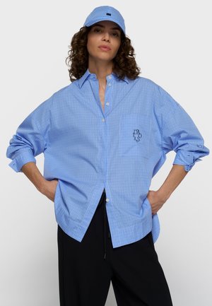 Vrouw met krullend haar draagt een lichtblauw geruit oversized overhemd en bijpassende pet, gecombineerd met een zwarte broek, handen in de zij, neutrale achtergrond.