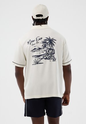 Άνδρας που φοράει κρεμ πουκάμισο και καπέλο, στην πλάτη του πουκαμίσου υπάρχει μαύρη παραλιακή σκηνή με φοίνικα, παράπηγμα ναυαγοσώστη και το κείμενο "New Era Since 1920."