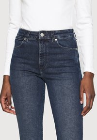 Donkerblauwe denim jeans met hoge taille, een gladde textuur, vijf zakken en een zilveren knoopsluiting aan de voorkant.