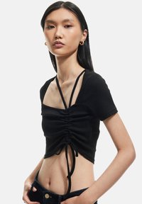 Ung kvinde iført en sort kortærmet rynket crop top med spaghetti stropper, blå jeans og guld hoop øreringe foran en hvid baggrund.