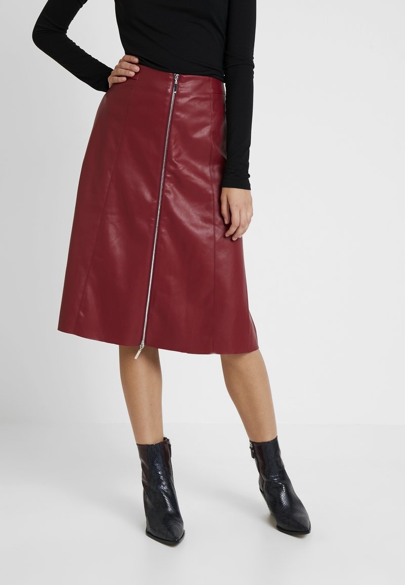 Jupe en simili cuir bordeaux avec une fermeture éclair sur le devant, longueur genou, texture lisse, associée à des bottines noires texturées.