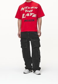 Camiseta roja de gran tamaño con el texto "NUNCA ES DEMASIADO TARDE" en crema y negro. Pantalones cargo negros con múltiples bolsillos. Zapatillas blancas.