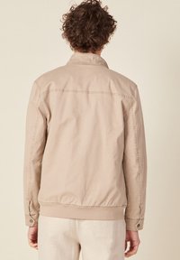 Beige Jacke mit Kragen, elastischem Bund und geknöpften Manschetten. Hergestellt aus weichem Stoff mit subtilen Nähten auf der Rückseite.
