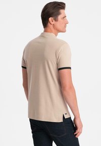Polo beige con maniche corte, bordo nero sulle maniche e dettagli con spacco laterale. Realizzato in tessuto morbido, design aderente e minimal branding.