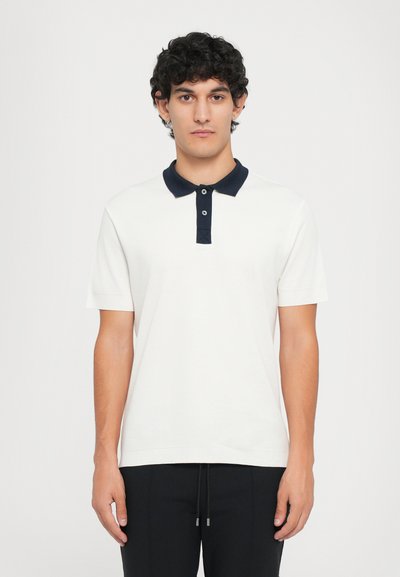 Weißes Poloshirt mit einem marineblauen Kragen, drei Knöpfen, kurzen Ärmeln und subtiler Textur, kombiniert mit schwarzen Jogginghosen mit Kordelzug.