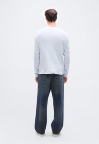 Άνδρας που στέκεται με την πλάτη γυρισμένη, φορώντας ανοιχτό γαλάζιο πουκάμισο με μακριά μανίκια, φαρδιά σκούρα μπλε τζιν και λευκά παπούτσια, πάνω σε απλό φωτεινό φόντο.