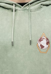 Sweat à capuche vert clair avec cordons de serrage et un graphique de globe marron et jaune sur la poitrine montrant les continents et les lignes de grille.