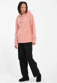 Pink hoodie made of soft fabric with a front pocket and drawstring hood, paired with black pants and black footwear. Simple design.