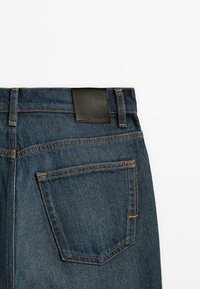 Jeans en denim foncé avec une texture lisse, présentant des coutures orange, une étiquette en cuir sur la taille et une poche arrière du côté droit.