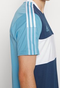 Blå och marinblå sporttröja med randigt mönster på ärmarna, tillverkad av lättviktsmaterial med rund hals och logodetalj.