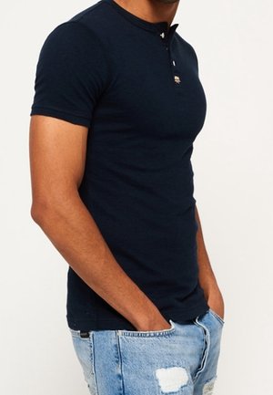 Mørkeblå, kortærmet, tætsiddende t-shirt med en lille tre-knaps placket. Bruges med lyseblå slidte denimjeans.