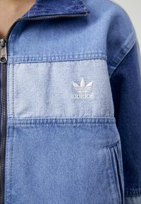 Jeansjacka i gradienttoner av blått med dragkedja och upprätt krage. Har broderad vit Adidas-logotyp på bröstfickan.