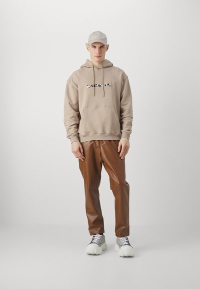 Sudadera marrón con texto gráfico, pantalones de imitación de cuero marrón y zapatillas blancas chunky. Una gorra gris claro completa el conjunto. Diseño limpio y minimalista.
