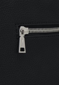 Črn teksturiran material s srebrnim zadrgo. Držalo zadrge ima kvadratno obliko z ikono klicaja. Poglej od blizu.