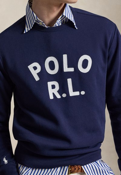 Homme portant un sweat-shirt bleu marine avec des lettres blanches « POLO R.L. » sur une chemise à rayures bleues et blanches avec col.