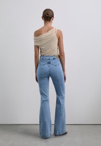 Vrouw met een bun, gekleed in een beige off-shoulder top en lichtblauwe high-waisted flared jeans, staande met het gezicht naar een effen grijze muur.