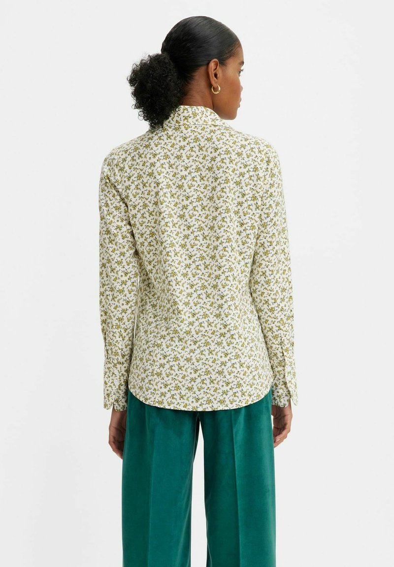 Langärmliges Shirt in Weiß mit grünem Blumenmuster, mit Kragen und abgerundetem Saum, kombiniert mit hellblauen, weit geschnittenen Hosen.
