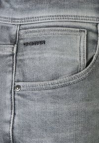 Jeans de denim gris claro con una textura deslavada. Presentan un logo negro de G-Star y un pequeño bolsillo con un botón a presión plateado.