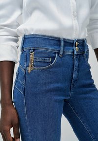 Dżinsy w ciemnoniebieskim kolorze z wysokim stanem, kontrastującymi złotymi elementami, przednimi kieszeniami oraz dekoracyjnym detalem w postaci frędzla z boku.