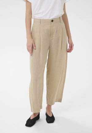 Personne portant un pantalon beige taille haute à jambes larges avec des rayures subtiles et des chaussures plates tissées noires, debout devant un fond blanc uni.