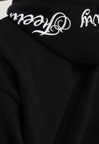 Felpa nera con una texture liscia, caratterizzata da un testo bianco in un font ornato lungo il bordo del cappuccio. Design semplice e classico.