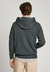 Sudadera gris hecha de tejido suave, con capucha ajustable y puños y dobladillo acanalados. Llevada sobre una camisa de cuadros, con las mangas visibles.