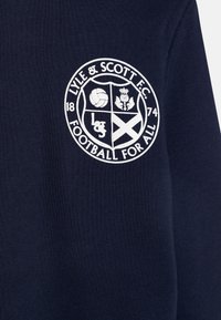 Sötétkék anyag, amelyen egy fehér hímzett kör alakú logó található. A logó tartalmaz egy focit, egy boglárt, a skóciai zászlót, valamint a "Lyle & Scott F.C." szöveget.