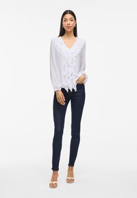 Hvit langermet bluse med volangdetaljer foran, V-hals og elastiske mansjetter, kombinert med mørke tettsittende jeans og åpne sandaler.