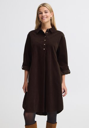 Woman wearing a dark brown buttoned dress with rolled-up sleeves, black tights, and brown boots against a plain light background.