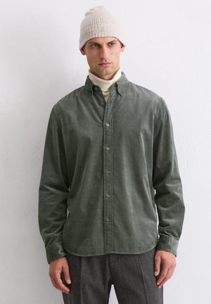 Grünes Cordhemd mit Knöpfen, kragenartig, strukturierte Oberfläche und slim fit, getragen über einem cremefarbenen Rollkragenpullover und kombiniert mit grauen Hosen.