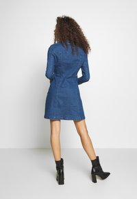 Sukienka jeansowa z długimi rękawami, dopasowaną talią i rozkloszowanym dołem; w głębokim niebieskim kolorze; z detalami przeszyć; zestawiona z czarnymi botkami na obcasie.