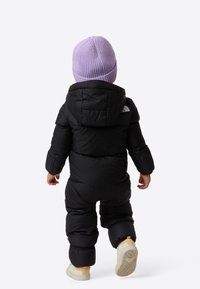 Black puffer snowsuit with a hood, featuring a white logo on the shoulder. Paired with a light purple knitted hat and beige boots.