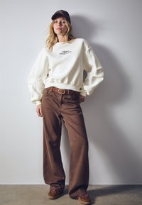 Felpa bianca oversize con polsini a costine, pantaloni marroni a gamba larga e scarpe marroni con lacci decorativi, su sfondo neutro.