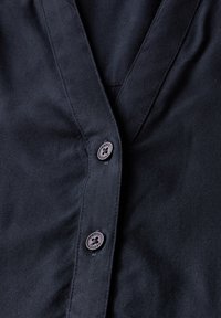 Camicia blu scuro con scollatura a V, tessuto liscio, cuciture visibili e un bottone rotondo con un design floreale vicino all'apertura.