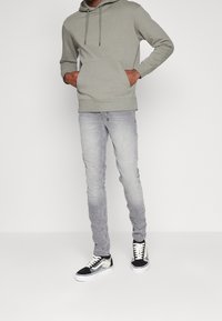 Sudadera gris clara con bolsillo frontal y cordones, combinada con jeans skinny gris desgastados y zapatillas negras con acentos blancos.