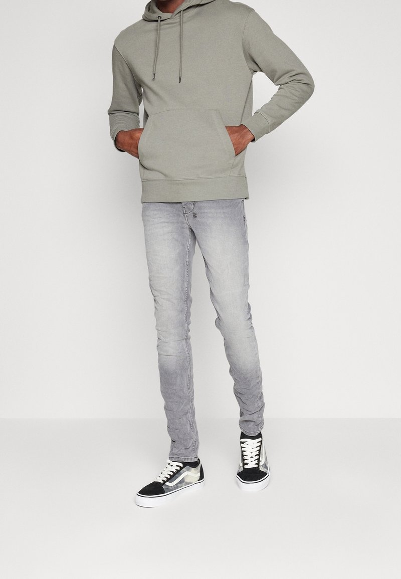 Sudadera gris clara con bolsillo frontal y cordones, combinada con jeans skinny gris desgastados y zapatillas negras con acentos blancos.