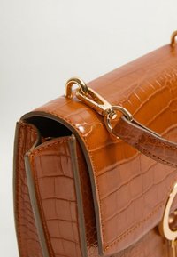 Brązowa torebka z tłoczonym wzorem aligatora, o ustrukturyzowanym kształcie, z złotymi okuciami i odpinanym paskiem. Charakteryzuje się kontrastowym przeszyciem i przednią klapą.