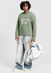 Olijfgroene sweater met textuur en de tekst "Relenity Atelier", gecombineerd met lichtblauwe versleten jeans en een unieke witte en grijze tas met textuur.