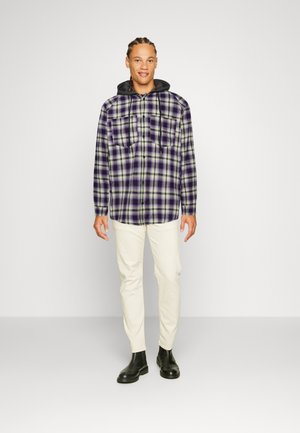 Skjorte i ternet flannel i lilla og grøn med to brystlommer, parret med kridhvide bukser og sorte ankle boots. Model står lige.