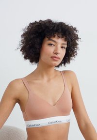 Bralette de Calvin Klein en suave tela beige, con un diseño de escote en V y una banda blanca con el logo. Textura suave con tirantes ajustables.