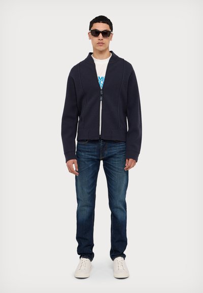 Navy spalvos zip-up striukė su aukštu apykakle, dėvima ant baltos grafikos marškinėlių, derinama su mėlynomis džinsomis ir baltais sportbačiais. Modelis turi juodus saulės akinius.
