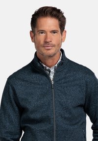 Dunkelblaues Zip-Jackett mit strukturiertem Strickoberfläche, ausgestattet mit einem Stehkragen und schlanker Passform, getragen über einem gemusterten Hemd.