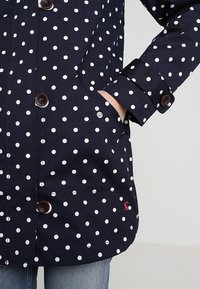 Mörkblå prickig kappa med vita prickar, bruna knappar och en röd logotyp på nederkanten. Tillverkad av ett slätt, strukturerat material.