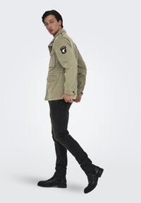Giacca di cotone khaki con design militare, dotata di una toppa sulla spalla, numerose tasche e chiusure a bottone. Abbinata a jeans neri e stivali.