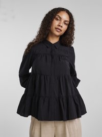 Ung kvinne med krøllete hår iført en svart lagdelt bluse og beige bukser, stående mot en ensfarget lys grå bakgrunn.