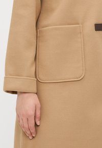 Bežinis vilnonis paltas su didele, stačiakampio formos kišene, turinčia švarią, siuvinėtą kraštą. Audinys turi lygų, šiek tiek tekstūruotą paviršių.