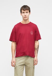 Νεαρός άνδρας με κοντά μαλλιά, φορώντας ένα χαλαρό κόκκινο T-shirt με λευκό κείμενο στο στήθος και μπεζ cargo παντελόνι, μπροστά σε απλό φόντο.