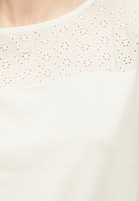 Λευκό τοπ με διαφανές μέρος στο πάνω μέρος με κεντητά λουλουδάτα μάτια, ντεκολτέ με κυματιστό άκρο που είναι ορατό στο πάνω μέρος του στήθους ενός ατόμου.