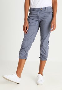 Grijze cropped broek met knoopsluiting, voorzien van zijzakken en geplooide details bij de zoom, gecombineerd met witte sneakers.