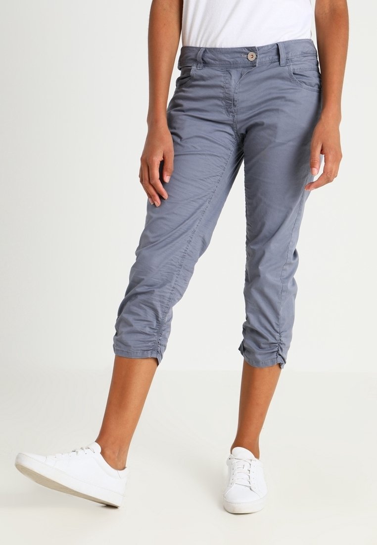 Grijze cropped broek met knoopsluiting, voorzien van zijzakken en geplooide details bij de zoom, gecombineerd met witte sneakers.